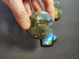 Hand holding a polished labradorite heart displaying blue and green iridescent flashes, with two similar stones visible in the background on a dark surface.