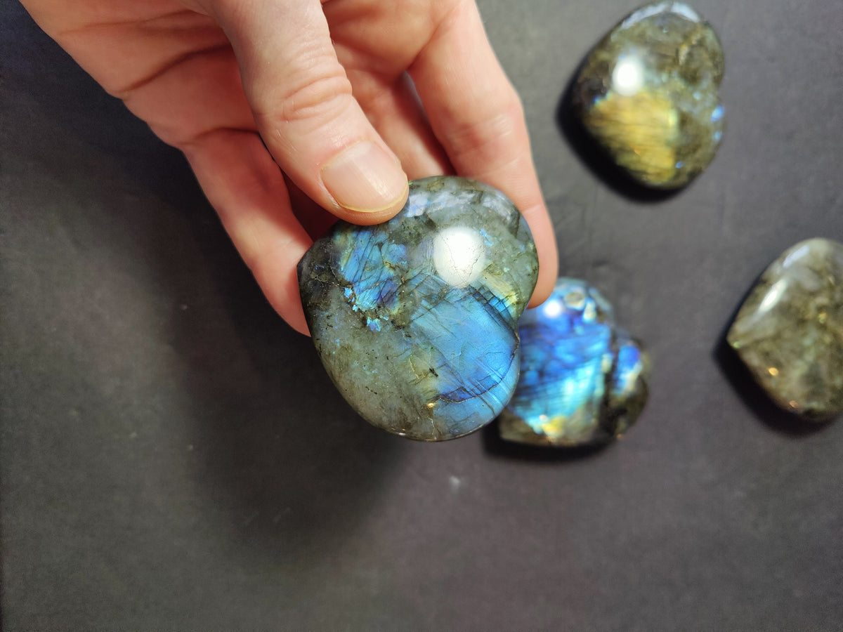 Hand holding a polished labradorite carved in a heart shape showing blue and green iridescent flashes, with three similar stones on a dark surface in the background.