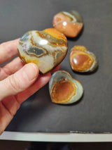 Hand holding a polished heart-shaped stone with multicolored swirls of orange, gray, and cream, resting above three similar stones on a dark background.
