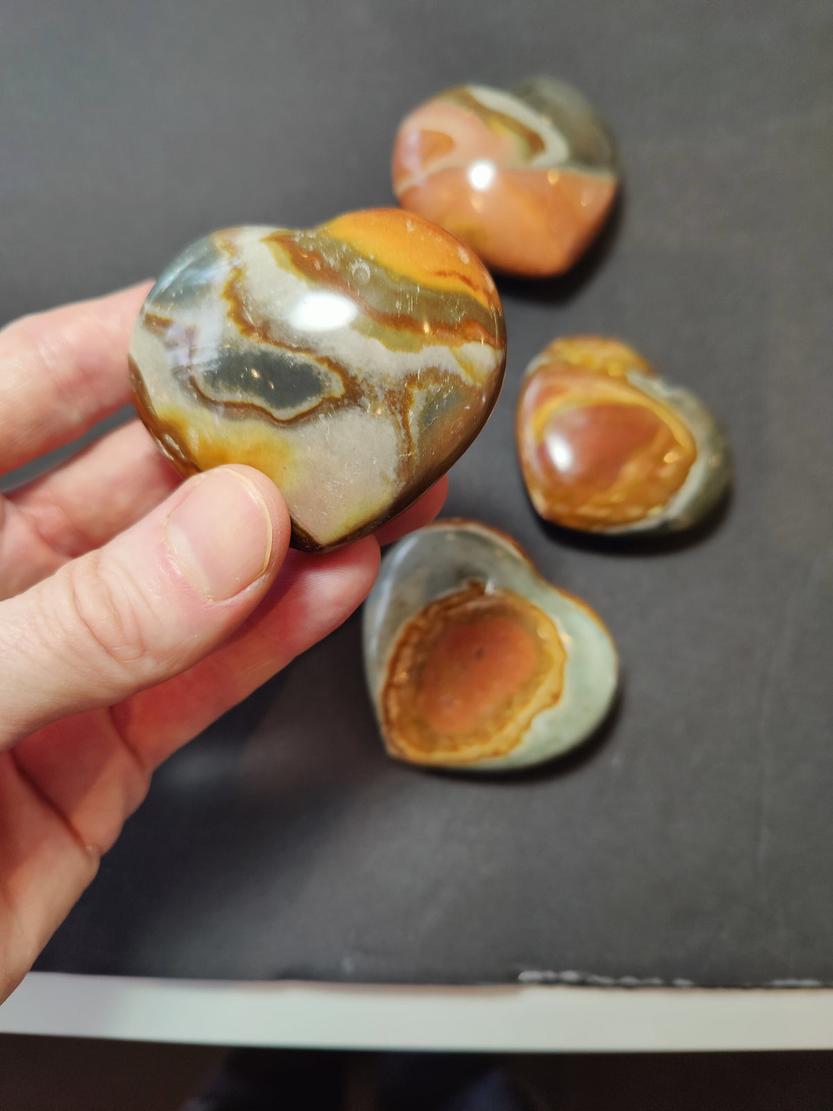 Hand holding a polished heart-shaped stone with multicolored swirls of orange, gray, and cream, resting above three similar stones on a dark background.
