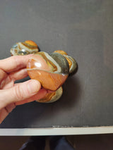 Hand holding a polished heart-shaped stone with brown, gray, and cream-yellow swirled patterns, with three similar stones blurred in the background on a dark surface.