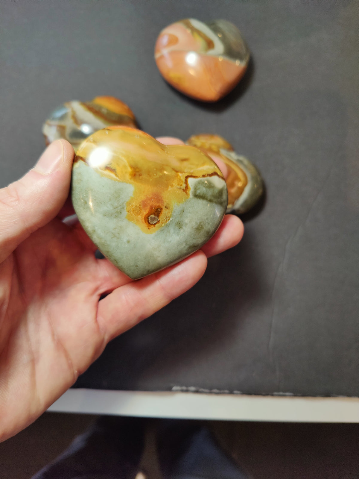Hand holding a polished heart-shaped stone with earthy green and orange-brown patterns, with three similar stones blurred in the background on a dark surface.