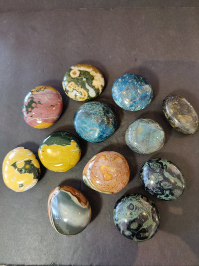 Assorted polished palmstones including Ocean Jasper, Blue Apatite, Kambaba Jasper, Polychrome Jasper, and Labradorite stones arranged on a wooden surface