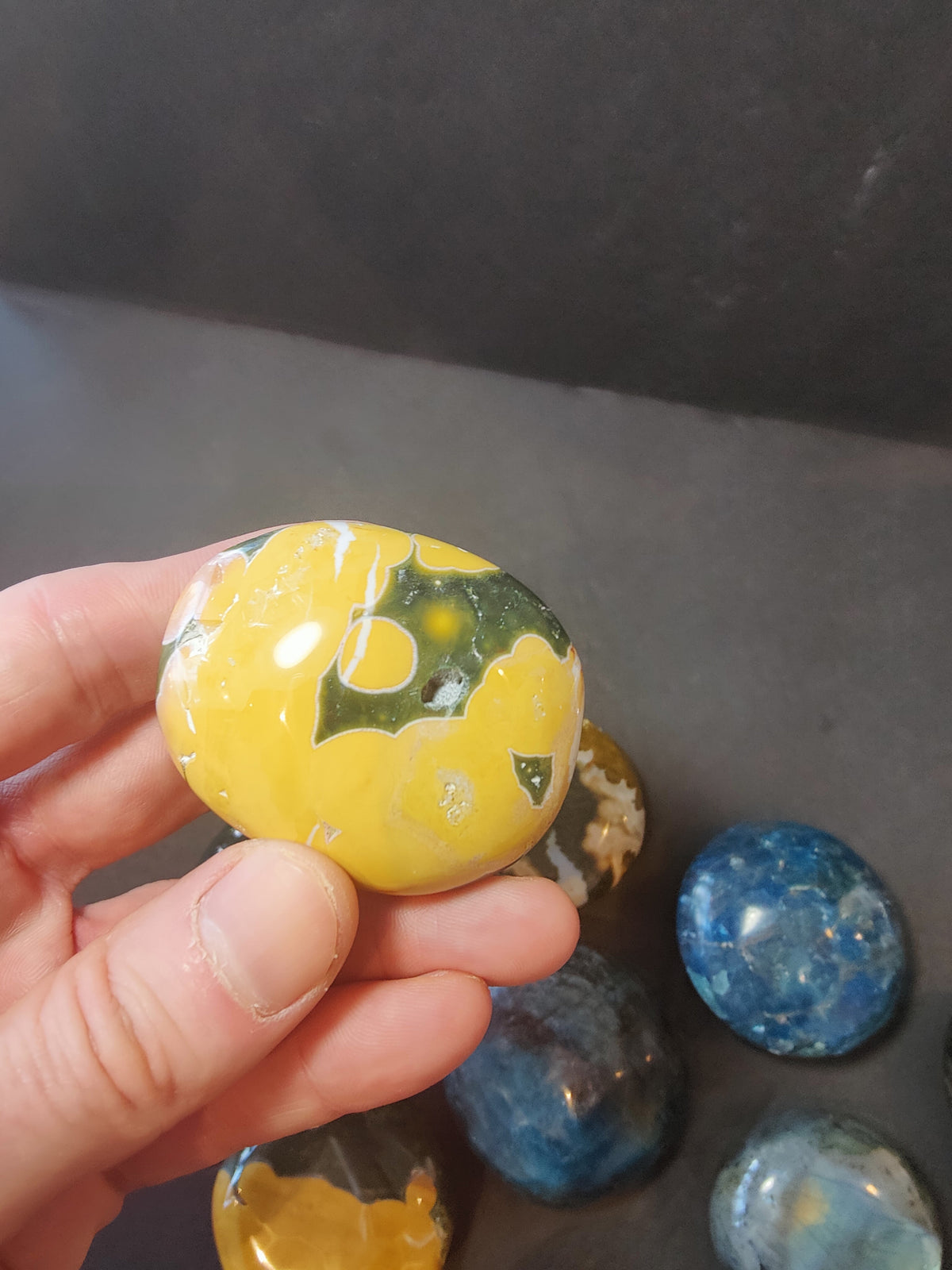 Hand holding a polished palmstone with yellow and dark green swirling patterns, other stones in shades of blue and brown visible in the background on a dark surface.