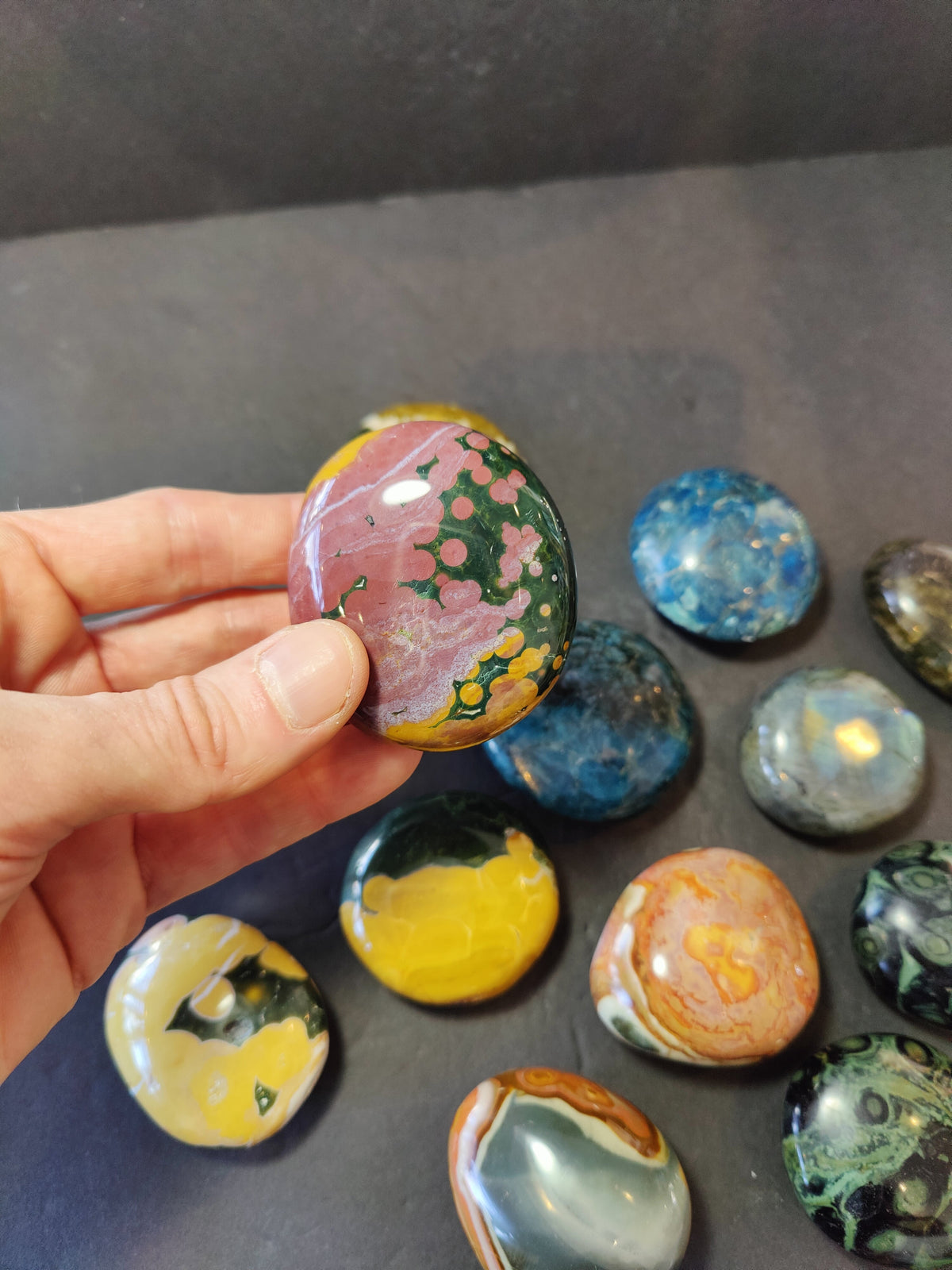 Hand holding a polished palm stone with green, pink, and mustard yellow patterns, surrounded by various smooth, multicolored polished stones on a dark surface.