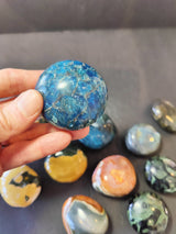 Hand holding a polished round blue palmstone with intricate lighter and darker blue patterns, surrounded by various other colorful polished stones on a dark surface.