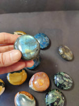 Hand holding a polished labradorite palmstone showing its blue-green iridescence, with various other polished stones in different colors and patterns scattered below on a dark surface.