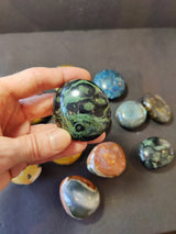 Hand holding a polished green and black patterned palmstone with other assorted polished stones in various colors blurred in the background on a dark surface.