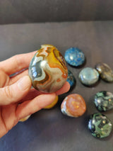 Close-up of a polished brown, orange, and white agate palmstone held between fingers, with assorted smooth blue, green, and multicolored stones in the blurred background.