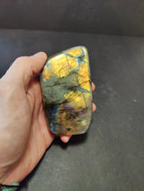 Hand holding a polished labradorite free form sculpture showing iridescent gold and blue flashes against a gray base with black veining.
