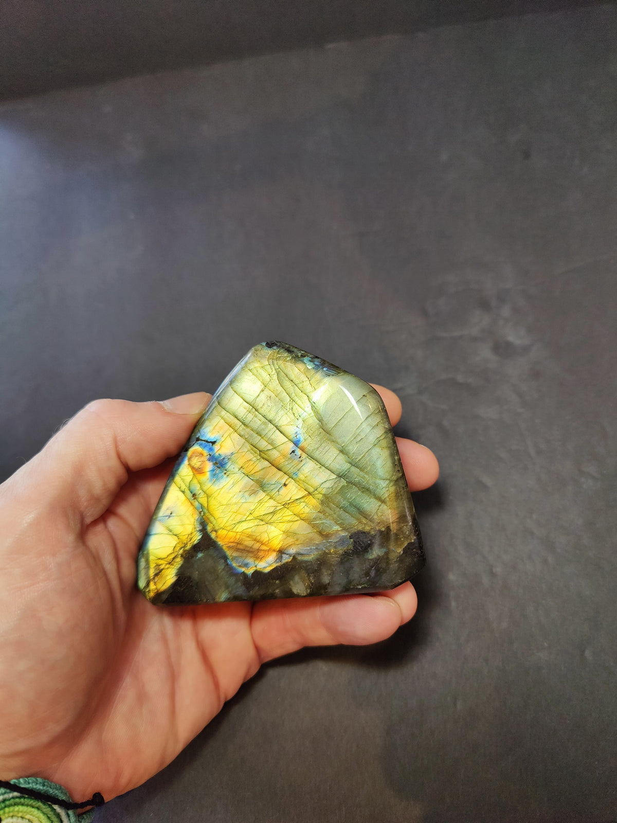 Hand holding a polished triangular labradorite stone exhibiting vibrant yellow and blue iridescent flashes against a dark, matte background.