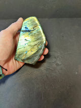 Hand holding a polished free-form labradorite stone showing iridescent green, blue, and gold flashes against a dark background.