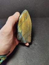 Hand holding a polished labradorite free form sculpture with golden striations and iridescent blue flashes on a black background.