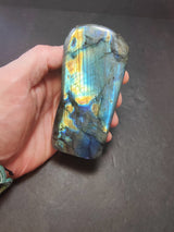 Hand holding a polished labradorite free form sculpture displaying iridescent blue, yellow, and orange flashes with natural gray and black veining.