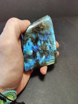 Hand holding a polished labradorite free form sculpture showcasing blue iridescent flashes against a dark gray stone matrix, with textured black veins.