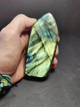 Hand holding a polished labradorite free form sculpture with iridescent blues and greens and dark veins, against a plain dark background.