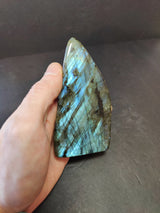 Hand holding a polished labradorite free-form sculpture with blue and green iridescent flashes and dark vein patterns on a gray background.