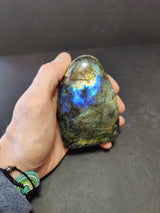 Hand holding a polished labradorite free form sculpture with blue and green iridescent flashes and smooth rounded top against a dark background.