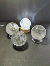 Four clear quartz polished spheres with natural inclusions, one elevated on a wooden stand and three on transparent bases, against a dark textured background.