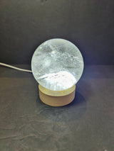 Clear quartz sphere with internal cracks and inclusions, polished and illuminated from below on a round wooden base against a dark background.