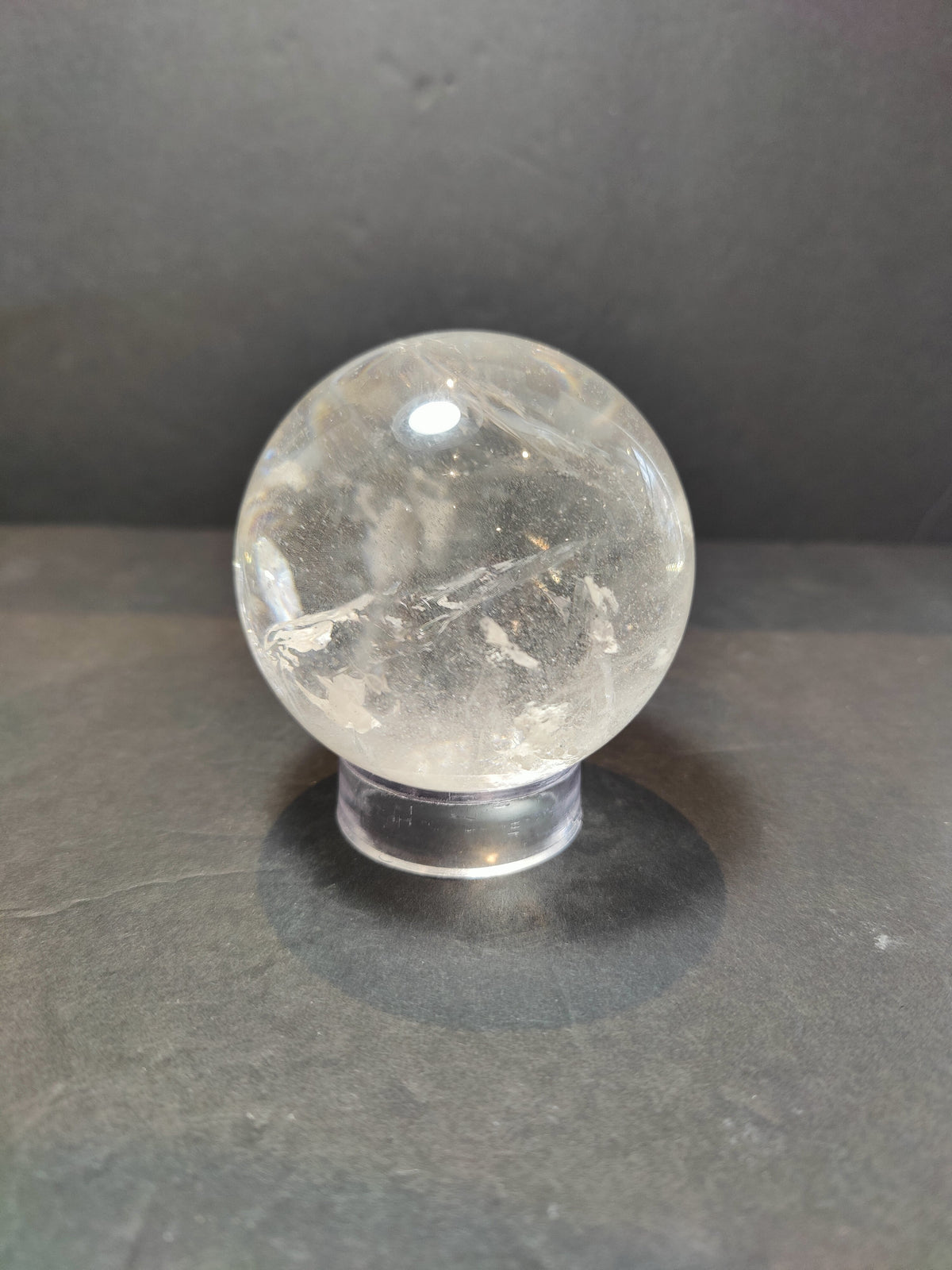 Polished clear quartz sphere with natural inclusions resting on a transparent round stand against a dark background.