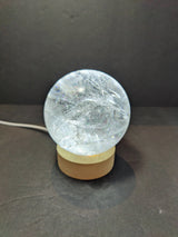 Transparent polished clear quartz sphere with internal natural inclusions, resting on a light wood round base against a dark background.