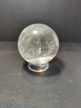 Polished clear quartz sphere with internal inclusions and reflective surface, resting on a small transparent stand against a dark background.
