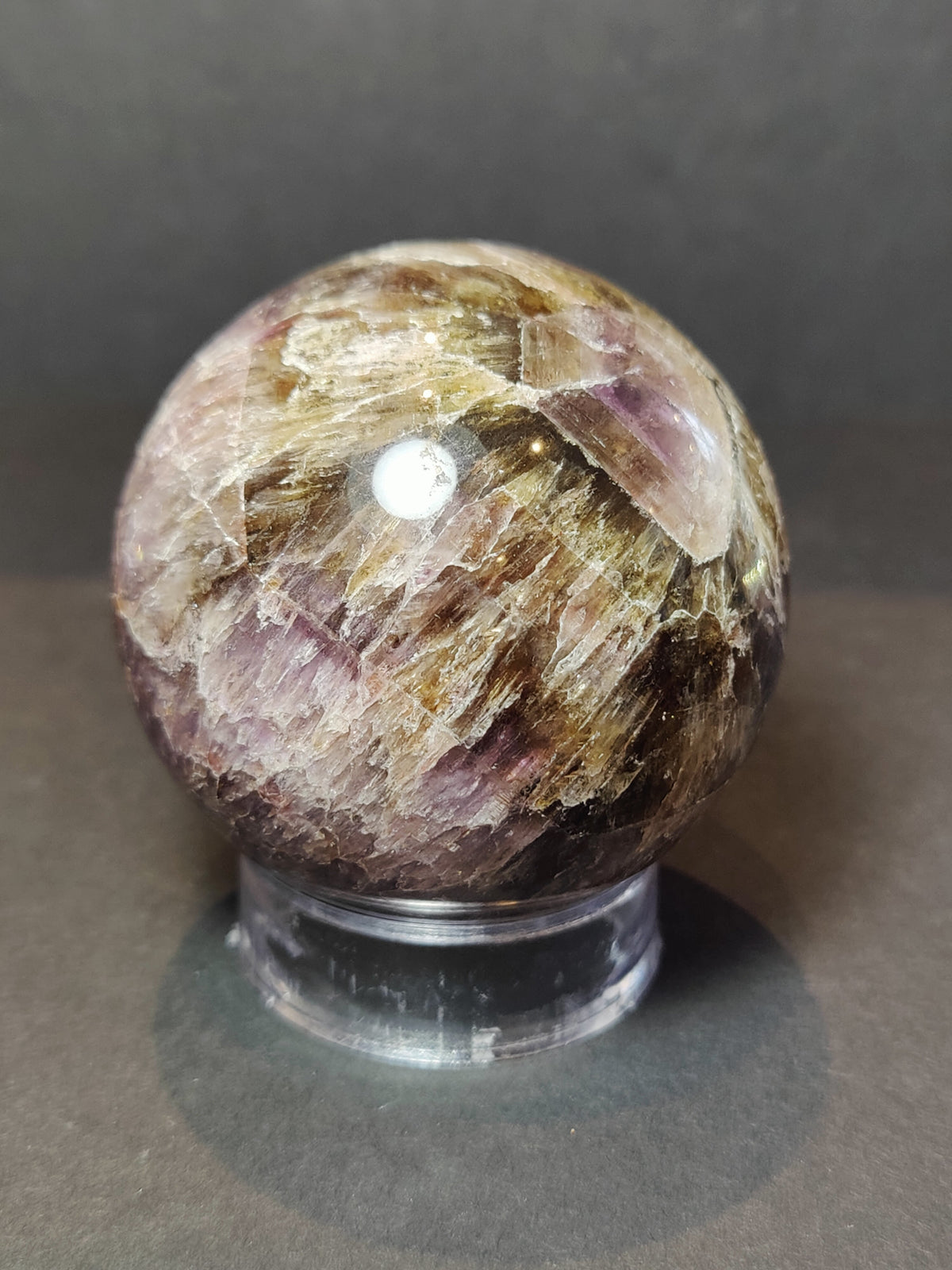Polished sphere with swirling patterns of purple and golden brown, resting on a clear round stand against a dark background.