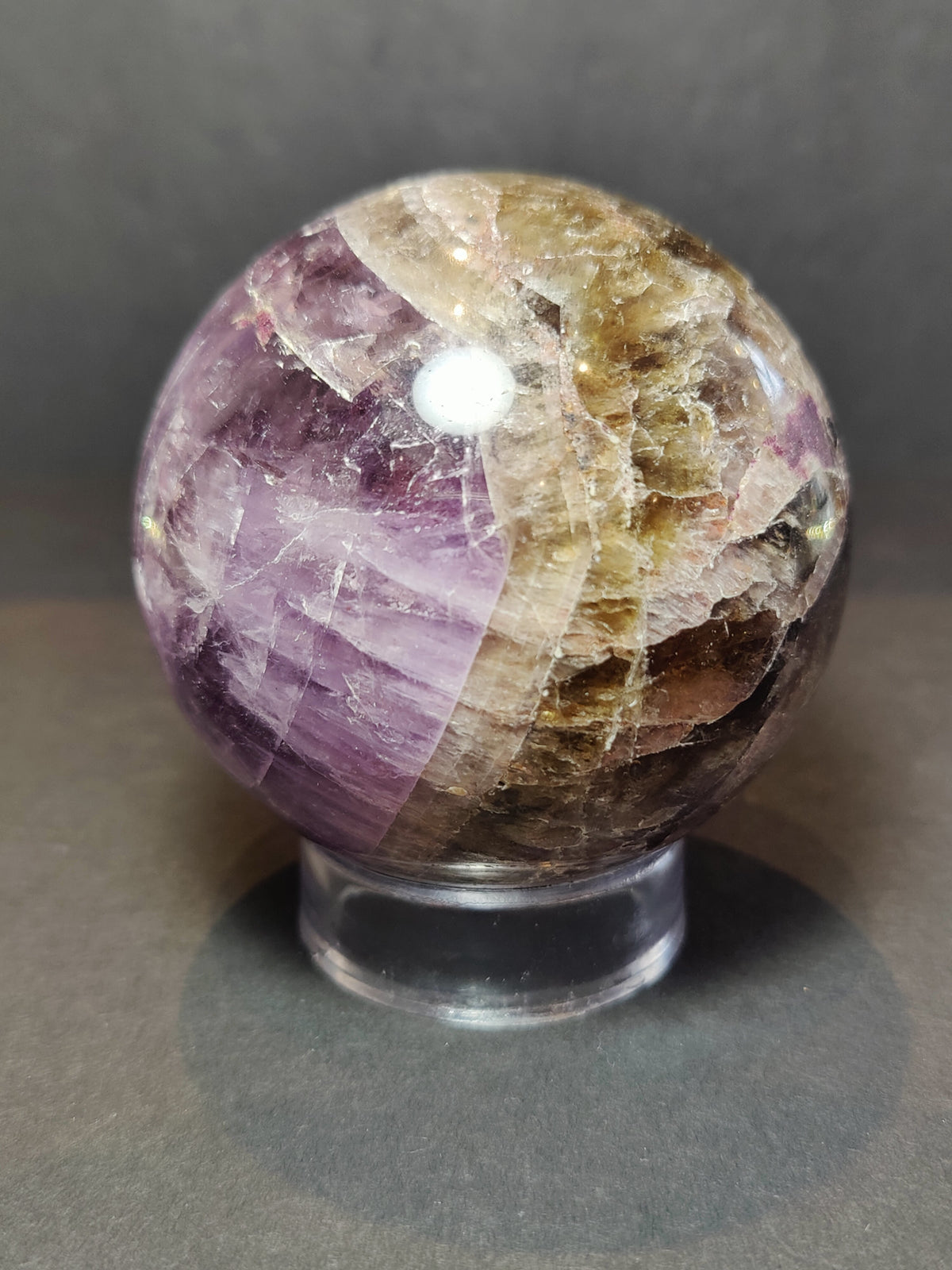 Polished spherical stone combining purple amethyst and tan cacoxenite with visible crystalline textures, resting on a clear stand against a gray background.