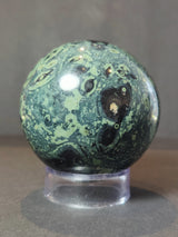 Polished Kambaba Jasper sphere with dark green and black swirling patterns, displayed on a clear cylindrical stand against a neutral background.