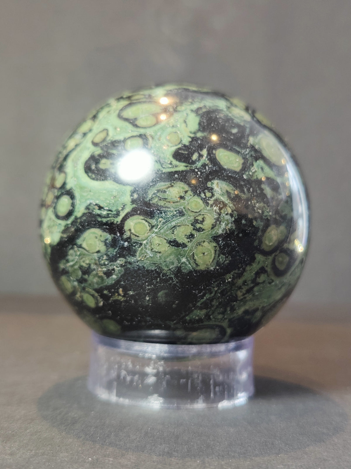 Polished Kambaba jasper sphere with dark green and black swirling circular patterns on a clear plastic stand against a gray background.