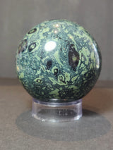 Polished Kambaba Jasper sphere with dark green and light green swirling patterns, displayed on a transparent round stand against a neutral background.
