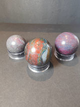 Three polished spheres displayed on clear stands, featuring shades of red, purple, and green with natural patterns and shimmering surface highlights.