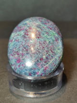 Oval polished stone with mixed blue, green, and ruby red hues, sitting on a clear round stand against a neutral background.