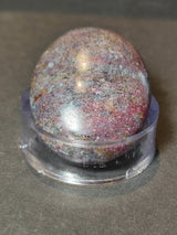 Oval polished stone with mixed reddish-purple and gray tones, resting on a clear circular plastic stand against a dark background.