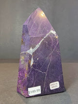 Polished purple purpurite obelisk tower with natural white veins and brown inclusions, displaying price and 'Made in Brazil' labels on a gray surface.