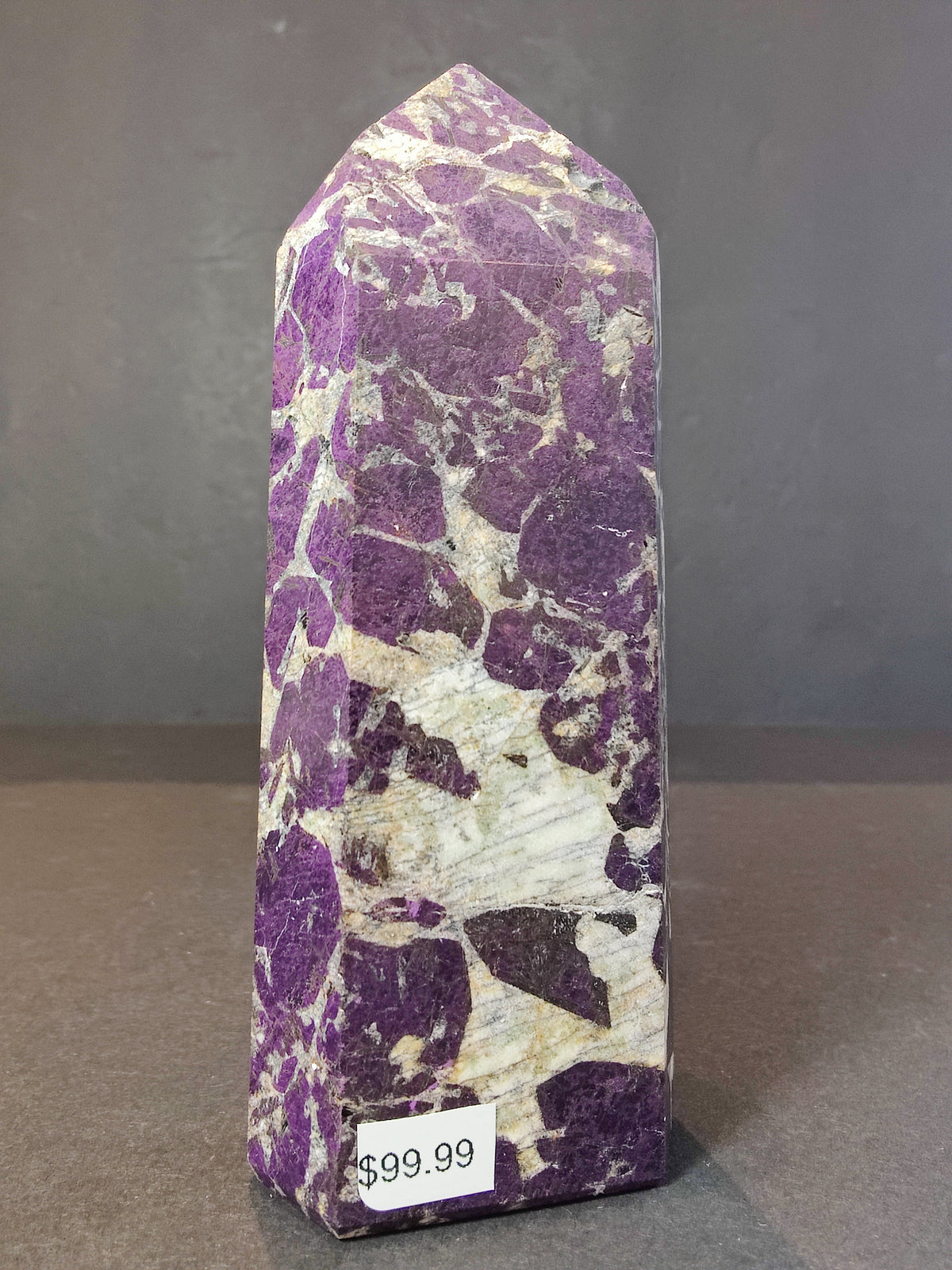 Polished obelisk-shaped purpurite stone with irregular purple and white marbled patterns standing upright against a gray background, labeled with a $99.99 price tag.