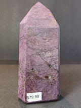 Polished purpurite obelisk tower sculpture with purple hues and natural veining, standing upright on a gray surface with a visible $79.99 price tag.