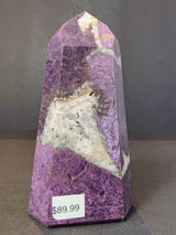 Polished purpurite obelisk tower sculpture with deep purple and white marbled patterns, standing on a dark surface with a price tag showing $89.99.