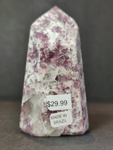 Polished lepidolite obelisk sculpture with purple and white crystalline patterns and price and origin labels on front against dark gray background.