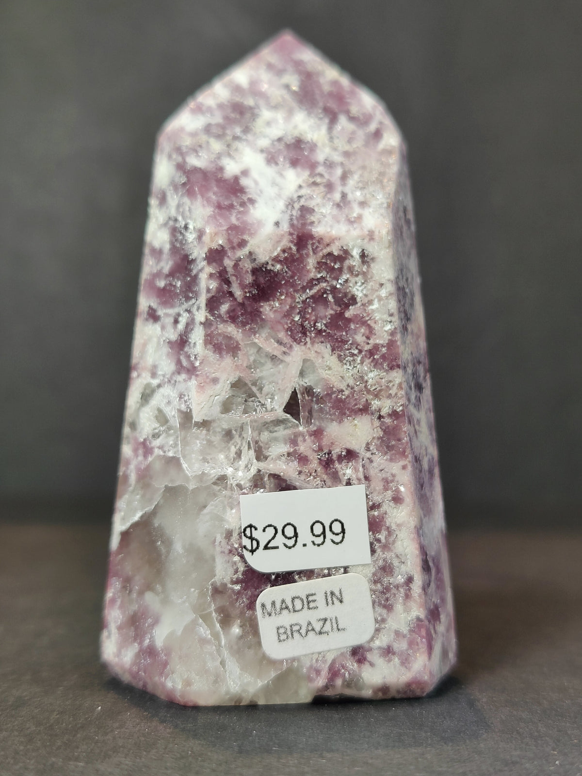 Polished lepidolite obelisk sculpture with purple and white crystalline patterns and price and origin labels on front against dark gray background.