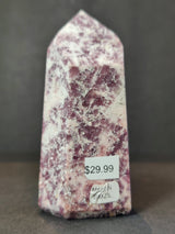 Polished lepidolite obelisk tower sculpture with mottled purple and white patterns, standing upright on a dark surface with price and origin labels attached.
