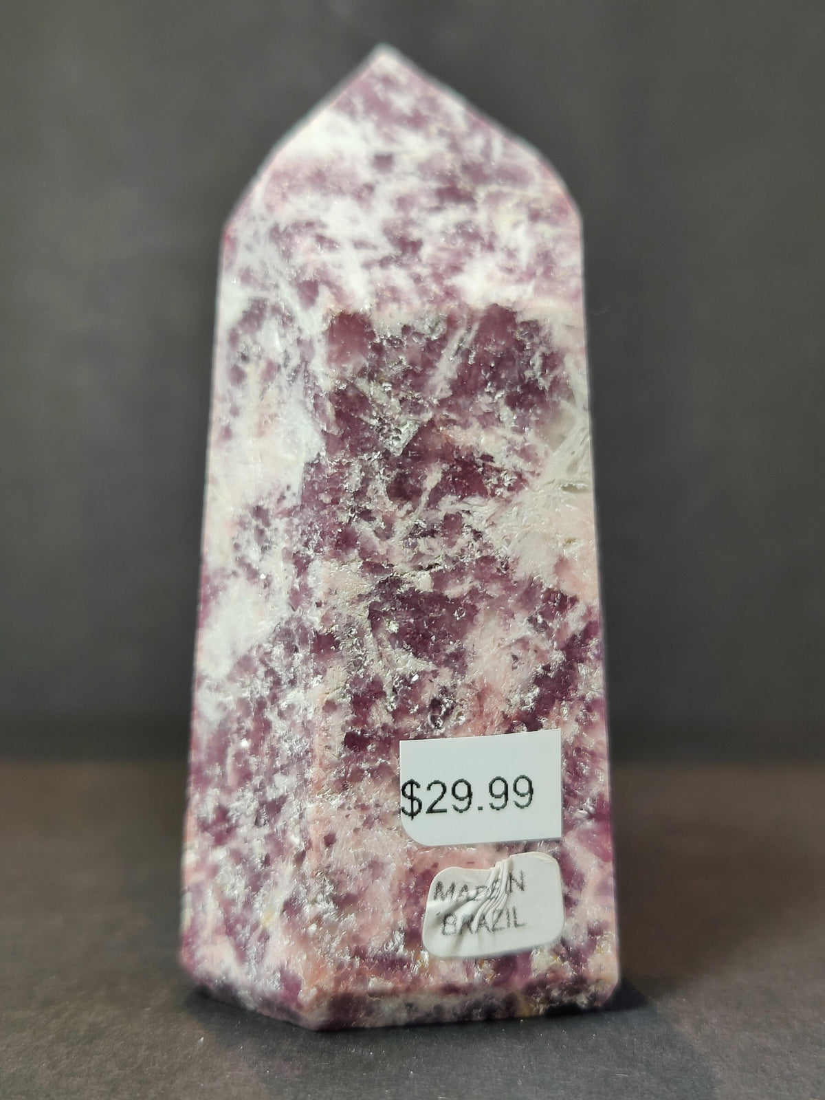 Polished lepidolite obelisk tower sculpture with mottled purple and white patterns, standing upright on a dark surface with price and origin labels attached.