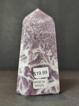 Polished lepidolite obelisk tower with purple and white marbled patterns standing against a dark gray background, labeled with a $19.99 price and 'Made in Brazil' sticker.