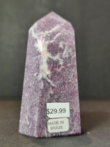 Polished lepidolite obelisk tower in purple with white marbling standing upright against a dark background, labeled with price and origin stickers.