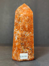 Polished sunstone obelisk tower sculpture in vibrant orange with specks of white and black, standing upright against a dark gray background.