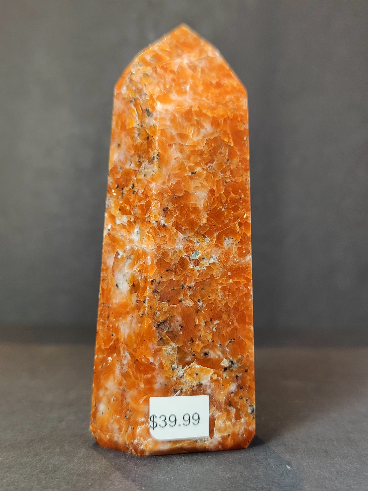 Polished sunstone obelisk tower sculpture in vibrant orange with specks of white and black, standing upright against a dark gray background.