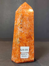 Polished obelisk tower sculpture made of orange sunstone with natural speckles, standing upright on a dark surface against a gray background.