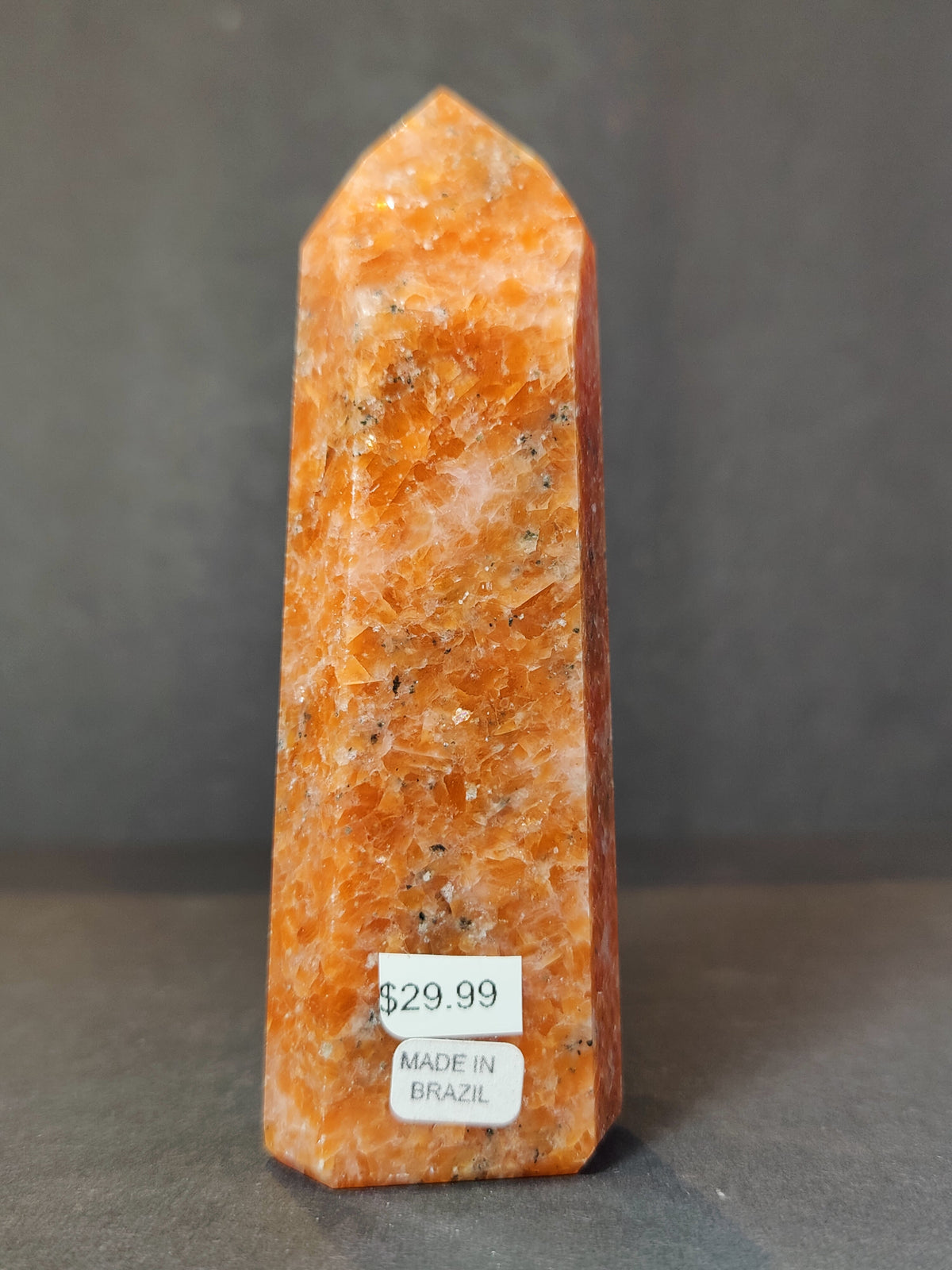 Polished orange sunstone obelisk with white and darker speckles, standing on a gray surface against a neutral background, tagged with price and origin labels.