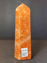 Tall polished obelisk sculpture with a warm orange and white mottled pattern and price stickers on its base, standing against a gray textured background.